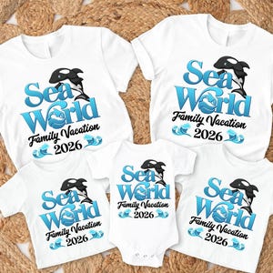 May include: White t-shirts and a onesie with a "Sea World Family Vacation 2026" design. The design features a blue and white wave, a black and white orca, and blue text. The shirts are short-sleeved.