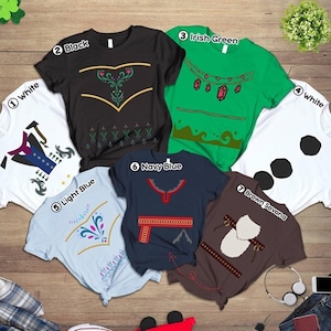 May include: Seven different coloured t-shirts with various Frozen character designs printed on them. The shirts are laid out on a wooden surface. The designs include Anna, Elsa, Kristoff, Olaf, and Sven.