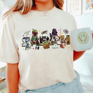 May include: Cream-colored t-shirt featuring the name "SARAH" in a colorful, Star Wars-themed design. The design includes characters like R2-D2, Boba Fett, Darth Vader, and Baby Yoda. A white mug with a smiley face is held in the image.