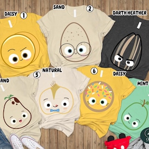 May include: Seven short-sleeve t-shirts in various colors including daisy yellow, sand, darth heather gray, and mint green. Each shirt features a unique cartoon character graphic with faces, including an egg, sunflower seed, potato, and donut.