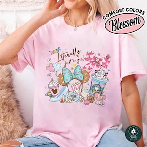 May include: A light pink t-shirt featuring a graphic print with the text "Literally just a girl" above illustrations of a blue glass slipper, a bow, a drink cup, a popcorn bucket, and a milkshake, all decorated with small hearts and stars.