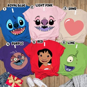 May include: A collection of six colorful t-shirts featuring cartoon character faces. The shirts are in royal blue, light pink, sand, purple, red, and lime green. Each shirt has a different character design, including a heart graphic.