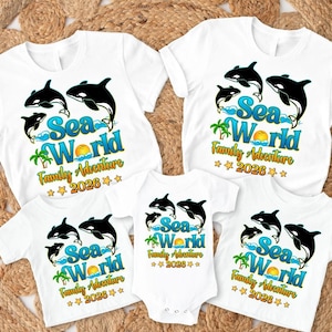 May include: White t-shirts and a onesie featuring a colorful "Sea World Family Adventure 2026" design with orcas, waves, a sun, and palm trees. The shirts are short-sleeved.