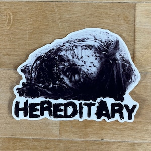 May include: A black and white sticker featuring a disturbing image of a face with the word "HEREDITARY" in bold, white letters. The sticker has a white border and is placed on a wooden surface.