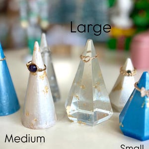 May include: Three ring display stands in different sizes: small, medium, and large. The small stand is blue, the medium stand is white with gold flecks, and the large stand is clear with gold flecks. Each stand has a ring on it.