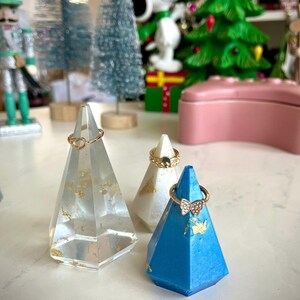 May include: Three geometric ring holders made of resin. The holders are in the shape of a pyramid and are decorated with gold leaf. The holders are in white, blue, and clear.