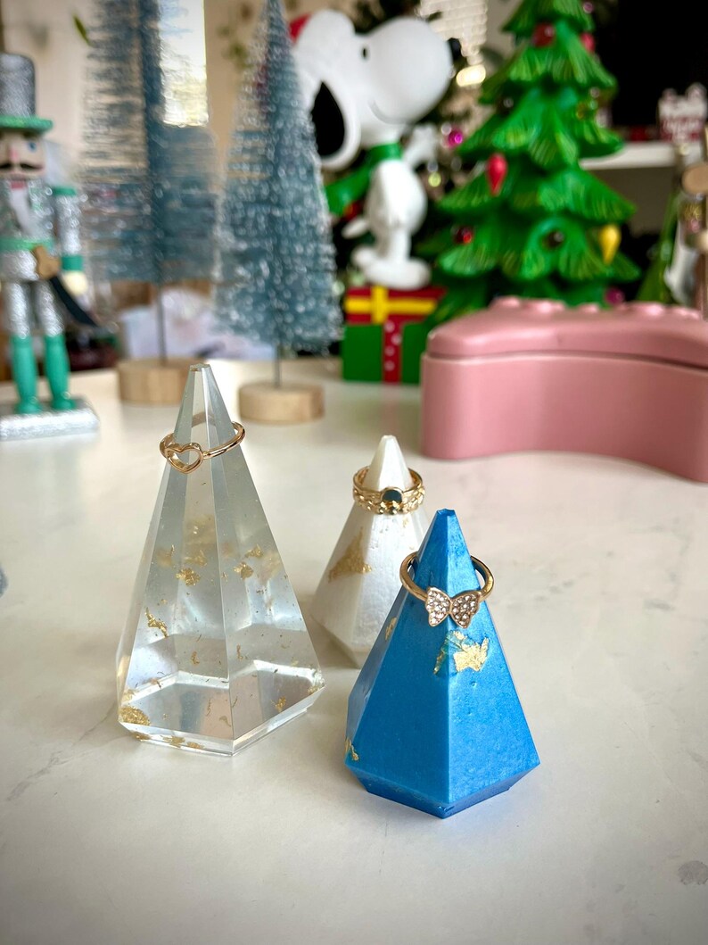May include: Three geometric ring holders made of resin. The holders are in the shape of a pyramid and are decorated with gold leaf. The holders are in white, blue, and clear.