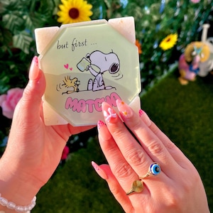 May include: A square, light green coaster with the text "but first... MATCHA" and a cartoon Snoopy and Woodstock design. The coaster is held in a person's hand, with a gold ring and a white pearl bracelet.