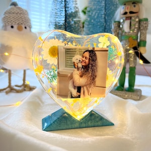 May include: A heart-shaped resin picture frame with a photo of a woman holding a small dog. The frame is decorated with dried flowers and lights. The frame is on a blue stand.