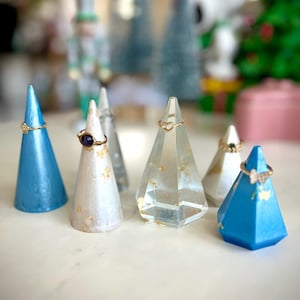 May include: Five ring display stands in different colors and materials. Two are blue, one is white with gold flecks, one is clear glass with gold flecks, and one is clear glass with a heart-shaped opening.