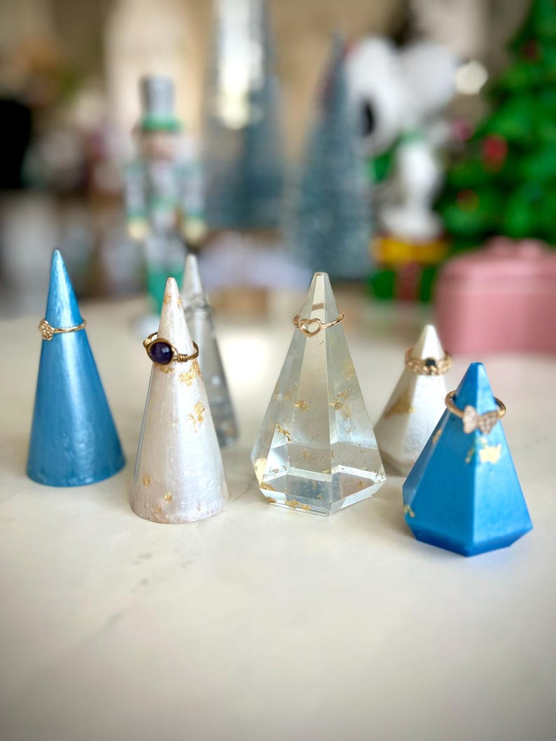 May include: Five ring display stands in different colors and materials. Two are blue, one is white with gold flecks, one is clear glass with gold flecks, and one is clear glass with a heart-shaped opening.