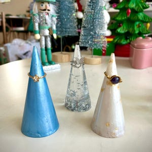 May include: Three ring holders, one blue, one clear with glitter, and one white with gold flecks. Each holder has a ring on it.