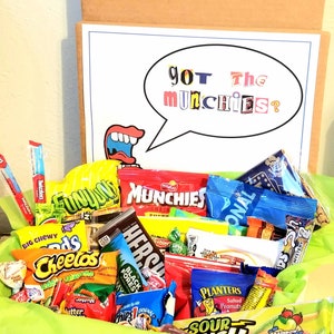 May include: A gift box filled with a variety of snacks, including chips, candy, and cookies. The box has a cardboard exterior and a green tissue paper lining. A sign inside the box reads "Got the Munchies?" in a speech bubble.