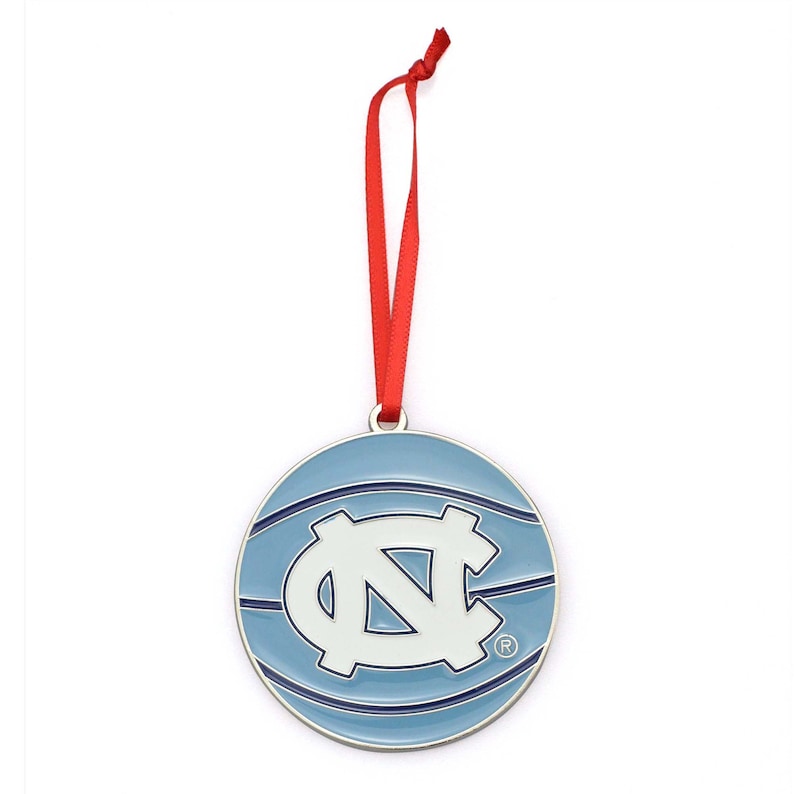 Puede incluir: Adorno redondo azul claro con borde plateado y cinta roja para colgar. Presenta el logotipo blanco de la Universidad de Carolina del Norte. El dise&ntilde;o recuerda a una pelota de baloncesto.