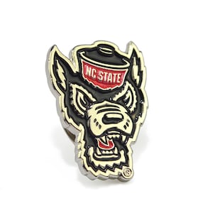 May include: A metal pin featuring the NC State Wolfpack logo. The pin has a black and red wolf head design with the words "NC STATE" on top. The pin has a silver border and is on a white background.