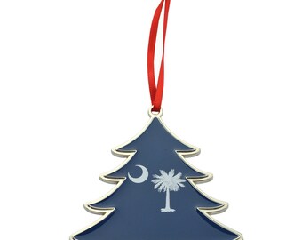 May include: A Christmas tree-shaped ornament with a navy blue background and a silver trim. The ornament features the South Carolina state flag design, including a white crescent moon and a palmetto tree. A red ribbon is attached for hanging.