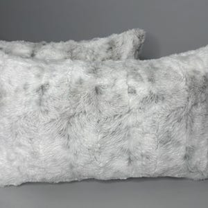 May include: Two rectangular decorative pillows with a faux fur texture. The pillows are primarily white with gray and black accents, creating a speckled appearance. The pillows are placed on a gray surface.