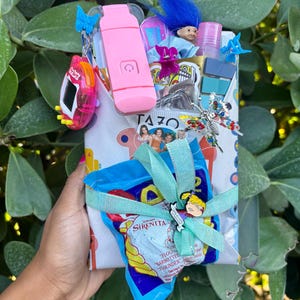 May include: A gift box wrapped in patterned paper, tied with a light blue ribbon and bow. The box contains a variety of items, including a pink electronic device, a blue-haired troll doll, and a small bag of snacks. Decorative charms and accessories are attached.