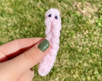 May include: A pink fuzzy worm plush toy with black eyes. The worm is being held in a hand with green nail polish.