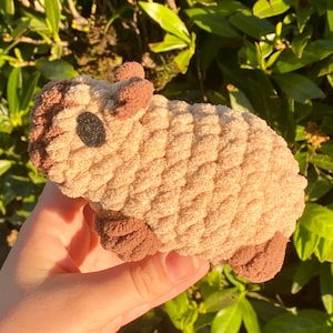May include: A handmade, plush toy in the shape of a small animal, possibly a sheep or lamb. The toy is primarily beige with brown accents on the ears, feet, and a small black eye. The plush material has a textured, looped appearance.