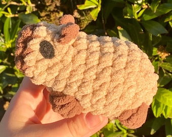 May include: A handmade, plush toy in the shape of a small animal, possibly a sheep or lamb. The toy is primarily beige with brown accents on the ears, feet, and a small black eye. The plush material has a textured, looped appearance.