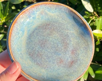 May include: A small, round ceramic dish with a speckled blue and brown glaze. The rim is a warm brown color, and the center has a mottled appearance. The dish is held in a hand, with green foliage in the background. This decorative dish is ideal for holding small items.