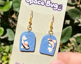 May include: A pair of handmade earrings featuring a blue and white koi fish design. The earrings are made of polymer clay and have gold hooks. The earrings are displayed on a card that says "The Space Bug" and "Handmade with Love".