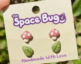May include: A set of three handmade polymer clay earrings featuring two red and white mushroom designs and one green leaf design. The earrings are packaged on a card with the text "The Space Bug" and "Handmade With Love".