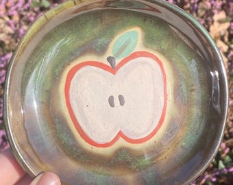 May include: A small, handmade ceramic dish with a painted apple design. The apple is white with a red outline, green leaf, and two black seeds. The dish has a green and brown glaze.
