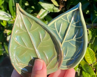 May include: Two leaf-shaped ceramic dishes in shades of green. The dishes have detailed veining and a glossy finish. The edges have a darker, contrasting color. The dishes are held in a hand against a background of green foliage.
