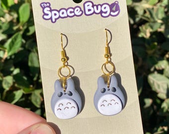 May include: A pair of gray and white Totoro dangle earrings with gold hooks. The earrings are handmade and feature the text "The Space Bug" and "Handmade With Love".