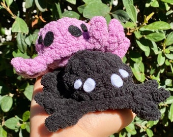 May include: Two plush toys, one black and one purple, with large white eyes. The black toy has a spider-like shape with eight legs. The purple toy has a rounded shape with two small ears.