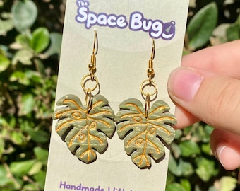 May include: A pair of green monstera leaf earrings with gold accents. The earrings are handmade and have a gold hook closure. The earrings are on a white card with the text "The Space Bug" and "Handmade With Love".