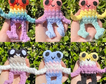 May include: Six crocheted frog plushies in various color combinations. Each frog has a different color pattern on its body, including rainbow, pink and purple, yellow and green, black and white, blue and pink, and purple and white.