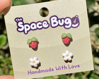 May include: A set of three handmade polymer clay earrings featuring a strawberry and two white flowers. The earrings are displayed on a card with the text "The Space Bug" and "Handmade With Love".
