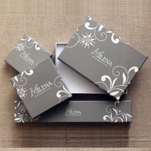 Pode incluir: Caixas de presente cinzentas com desenhos florais e redemoinhos brancos. As caixas, de v&aacute;rios tamanhos, est&atilde;o dispostas sobre uma superf&iacute;cie neutra. Algumas caixas est&atilde;o abertas, revelando um interior branco. Trazem o nome "Milena".