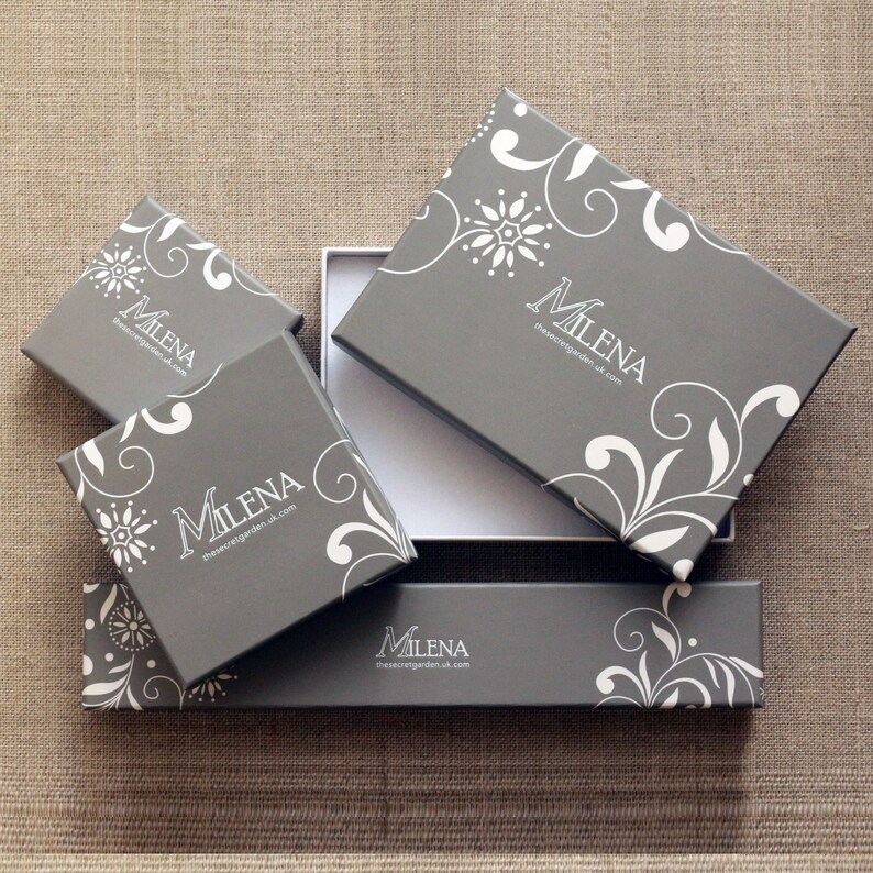 Pode incluir: Caixas de presente cinzentas com desenhos florais e redemoinhos brancos. As caixas, de v&aacute;rios tamanhos, est&atilde;o dispostas sobre uma superf&iacute;cie neutra. Algumas caixas est&atilde;o abertas, revelando um interior branco. Trazem o nome "Milena".