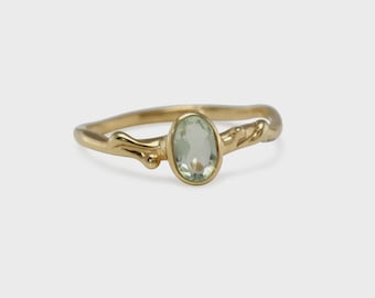 Puede incluir: Un anillo de oro con una piedra preciosa ovalada de color verde pálido. La banda del anillo tiene un diseño orgánico único con detalles texturizados. La piedra preciosa está engastada en un engaste sencillo, que resalta su color y forma naturales.