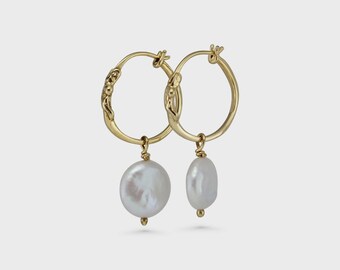 May include: A pair of gold-colored hoop earrings with a textured design. Each earring features a large, white, oval-shaped pearl pendant. The earrings are displayed against a plain, light background.