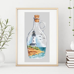 Puede incluir: Un diseño de punto de cruz enmarcado de un faro en una botella de vidrio. La botella es transparente y tiene un tapón de corcho. El faro es blanco con un techo negro y una linterna negra. La escena incluye una pequeña playa, agua azul y algunas aves volando por encima.