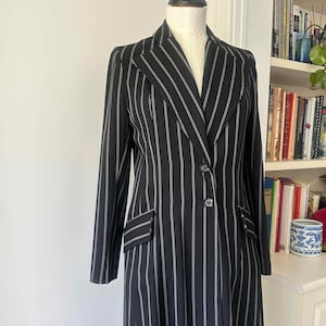Può includere: Un cappotto nero a gessati bianchi, esposto su un manichino. Il cappotto lungo presenta un colletto a revers, due tasche anteriori e un design doppiopetto con due bottoni. Le strisce verticali creano un effetto snellente. Il cappotto sembra essere realizzato in tessuto leggero.