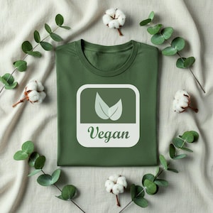 May include: A dark olive green t-shirt featuring a square graphic with rounded corners. The graphic displays two light green leaves above the word Vegan written in a cursive script. The design is centred on the front of the garment.