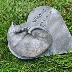 May include: A gray heart-shaped stone memorial with a sleeping cat sculpture. The stone has the inscription "Winston Love and miss you more than words".