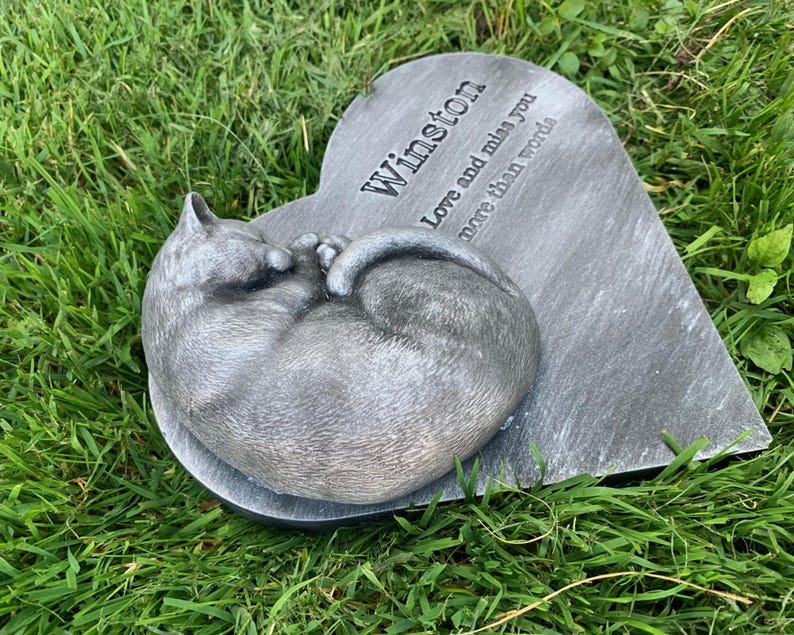 May include: A gray heart-shaped stone memorial with a sleeping cat sculpture. The stone has the inscription "Winston Love and miss you more than words".
