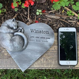 May include: A gray heart-shaped stone memorial with a sleeping cat sculpture. The stone has the inscription "Winston I love and miss you more than words".