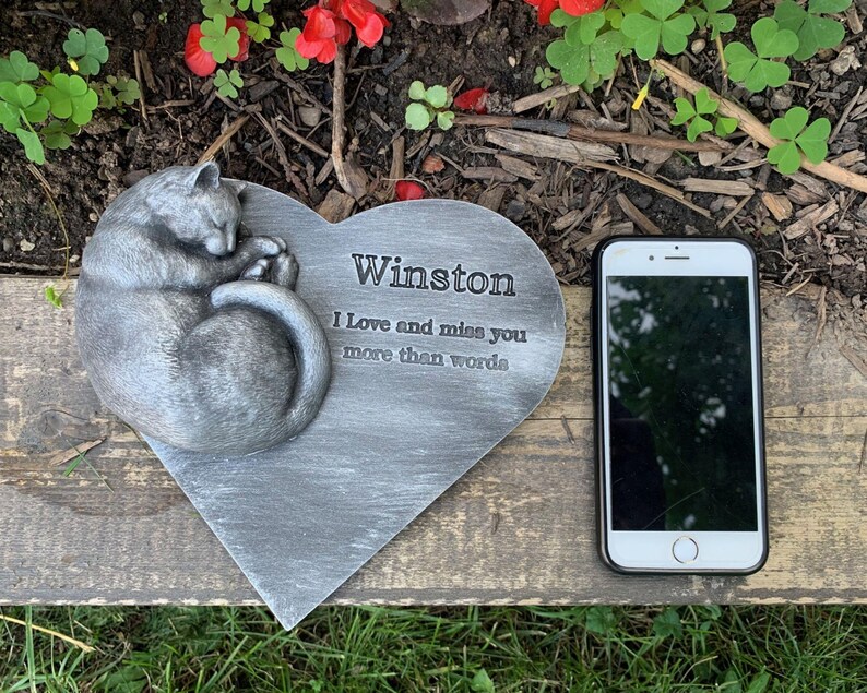 May include: A gray heart-shaped stone memorial with a sleeping cat sculpture. The stone has the inscription "Winston I love and miss you more than words".
