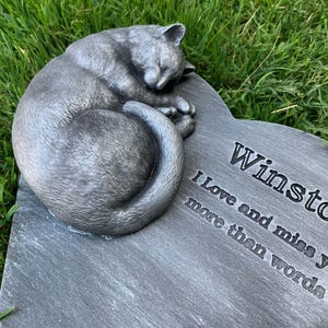 May include: A gray stone heart-shaped memorial with a sleeping cat sculpture. The stone is engraved with the text "Winston I love and miss you more than words".