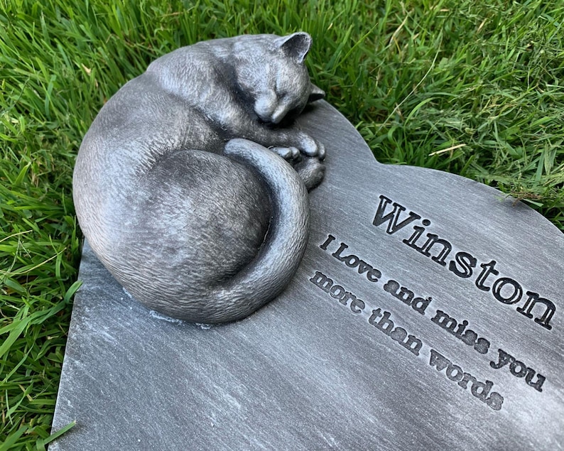 May include: A gray stone heart-shaped memorial with a sleeping cat sculpture. The stone is engraved with the text "Winston I love and miss you more than words".
