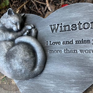 May include: A gray heart-shaped stone memorial with a sleeping cat sculpture. The stone has the inscription "Winston I love and miss you more than words."