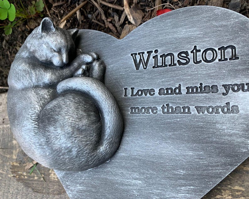 May include: A gray heart-shaped stone memorial with a sleeping cat sculpture. The stone has the inscription "Winston I love and miss you more than words."